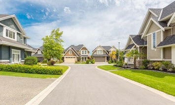 Wide paved street lined with modern suburban houses featuring manicured lawns, trees, and clear skies in the background.