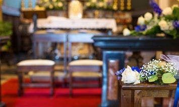 Flowers rest on a wooden pew adorned with white and blue ribbons, within a decorated church featuring an altar, chairs, and a red carpet leading down the aisle in the background.