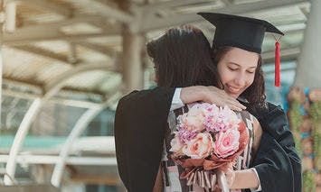 A graduate wearing a cap and gown hugs another person tightly, holding a bouquet of pink flowers in an outdoor setting with structural beams and people in the background.