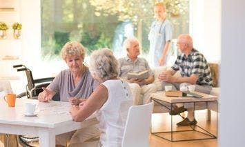 Elderly individuals converse at a bright, airy senior living facility. Two women sit and talk at a table, two men sit nearby, while a caregiver stands in the background.