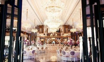 Crystal chandeliers illuminate an opulent ballroom set for an event with elegantly arranged round tables and floral centerpieces, viewed through open double doors.